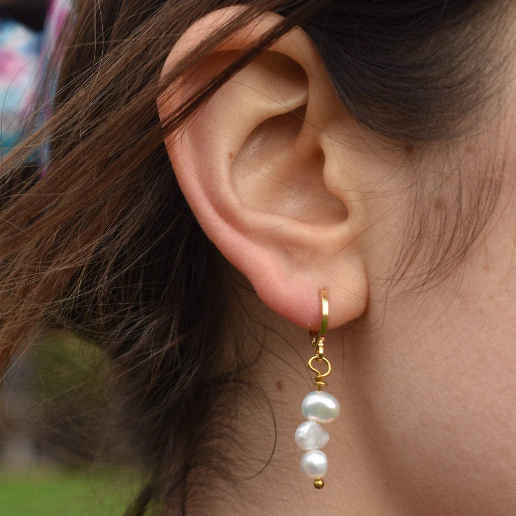 Triple irregular white freshwater pearls dangle from gold-plated brass ear huggies, worn on the ear.